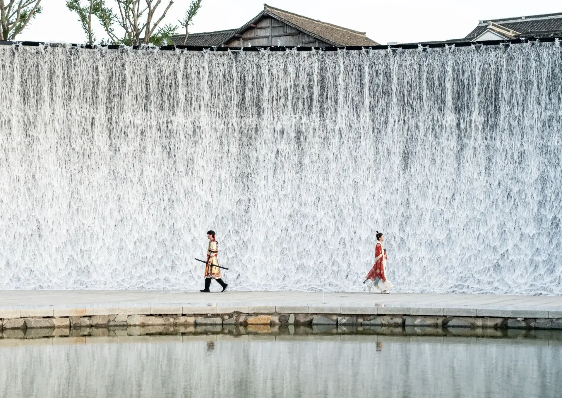 Two people in ancient costumes walk in front of Yanguan waterfall tide wall with water reflection.