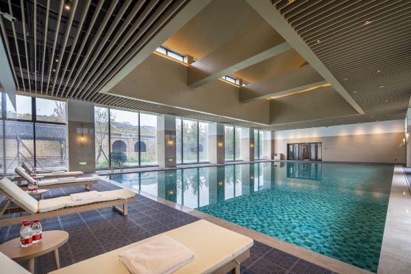 An indoor swimming pool with soft lighting, lounge chairs, and a modern ceiling design creating a relaxing atmosphere.