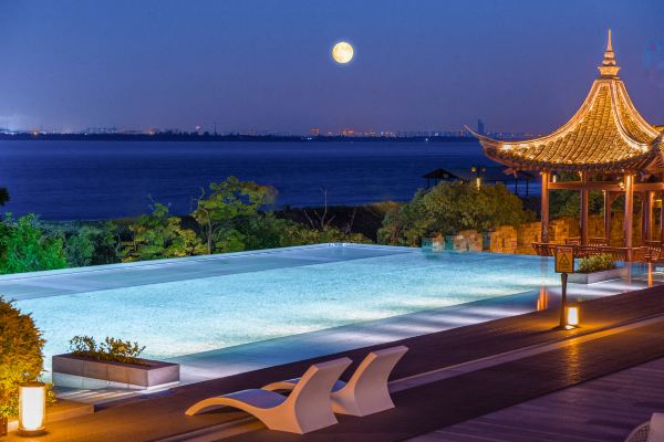An outdoor infinity pool overlooking the sea at dusk, with a pavilion and warm lights reflecting on the water.