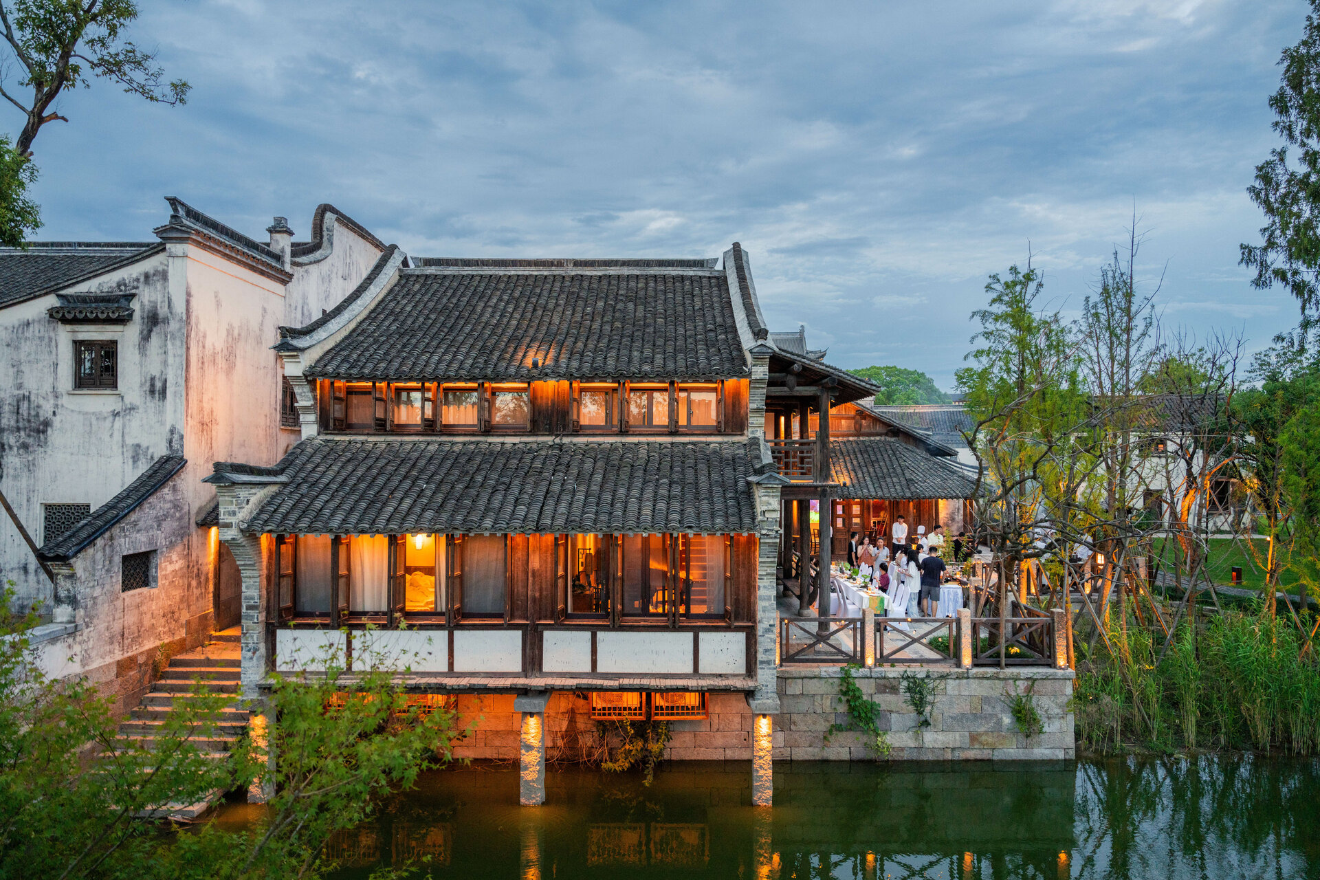 Twilight shot of traditional Puyuan waterside residence with guests dining beside canal.