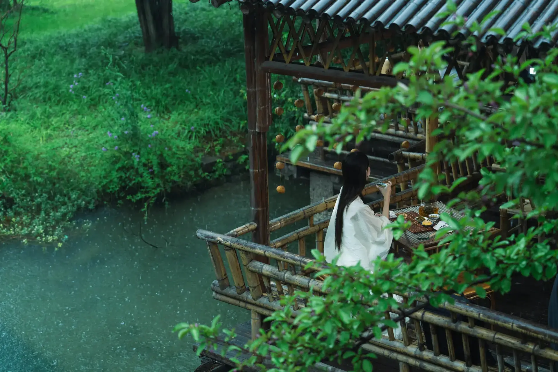 Girl in Hanfu tasting tea on waterside bamboo veranda in rain.