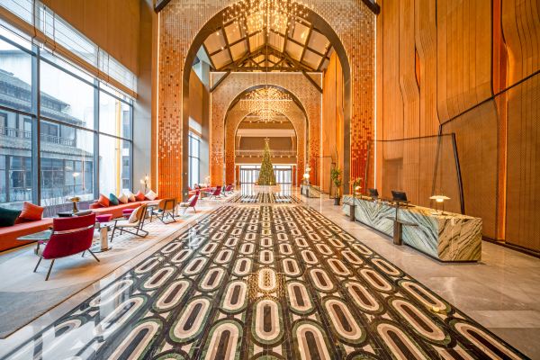A grand hotel lobby featuring high ceilings, golden accents, and a long patterned carpet leading through the space.