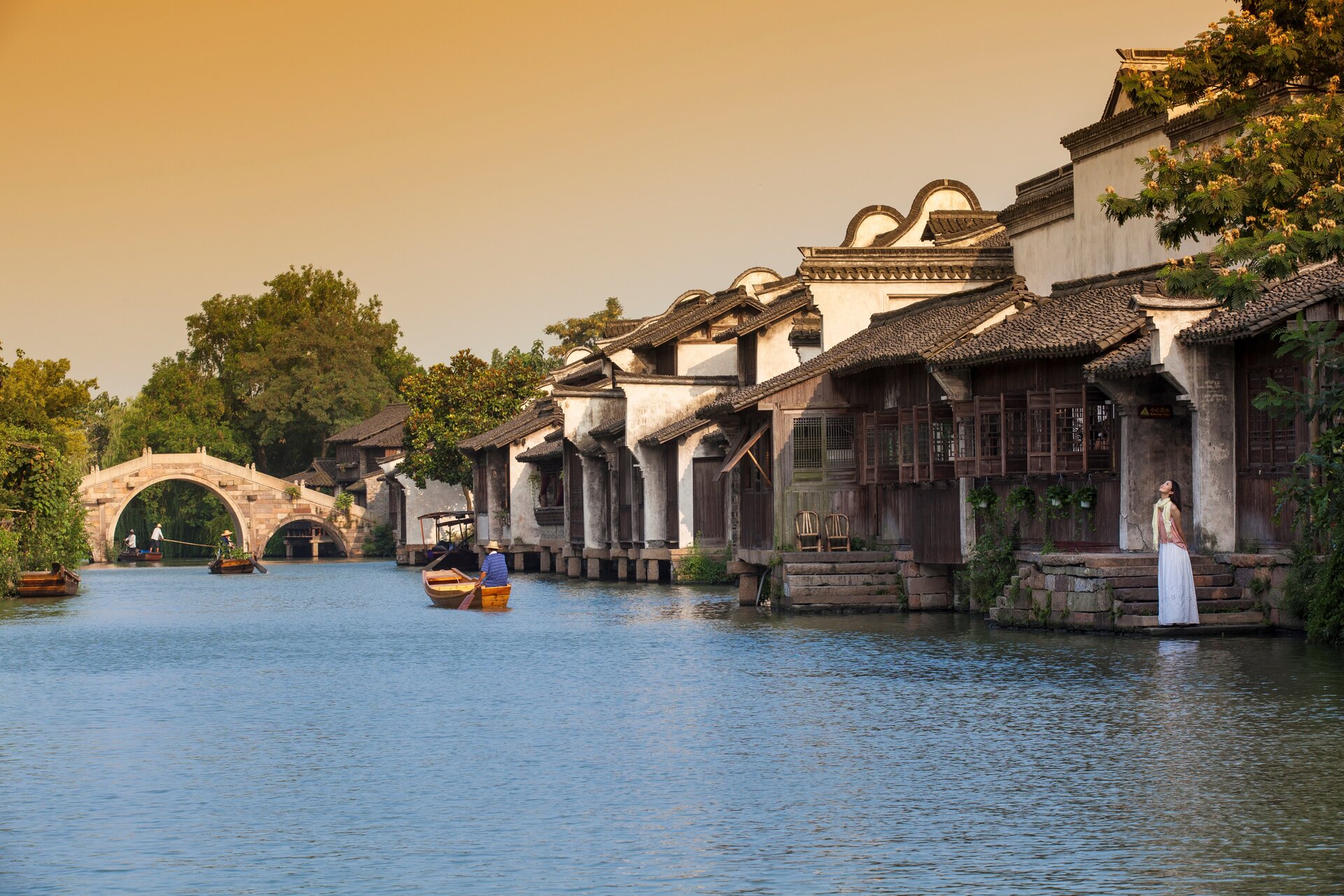 Riverside Huizhou-style wooden houses, stone arch bridge and rowing boats, lady in long white dress standing by water steps under warm sunset sky.