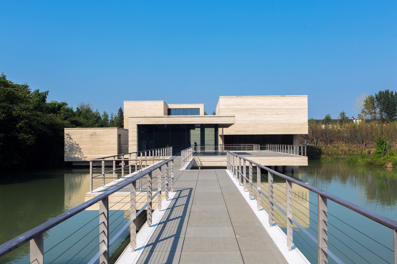 Modern minimalist museum building extending over water, connected by a long walkway, with clean geometric architecture and calm surroundings.