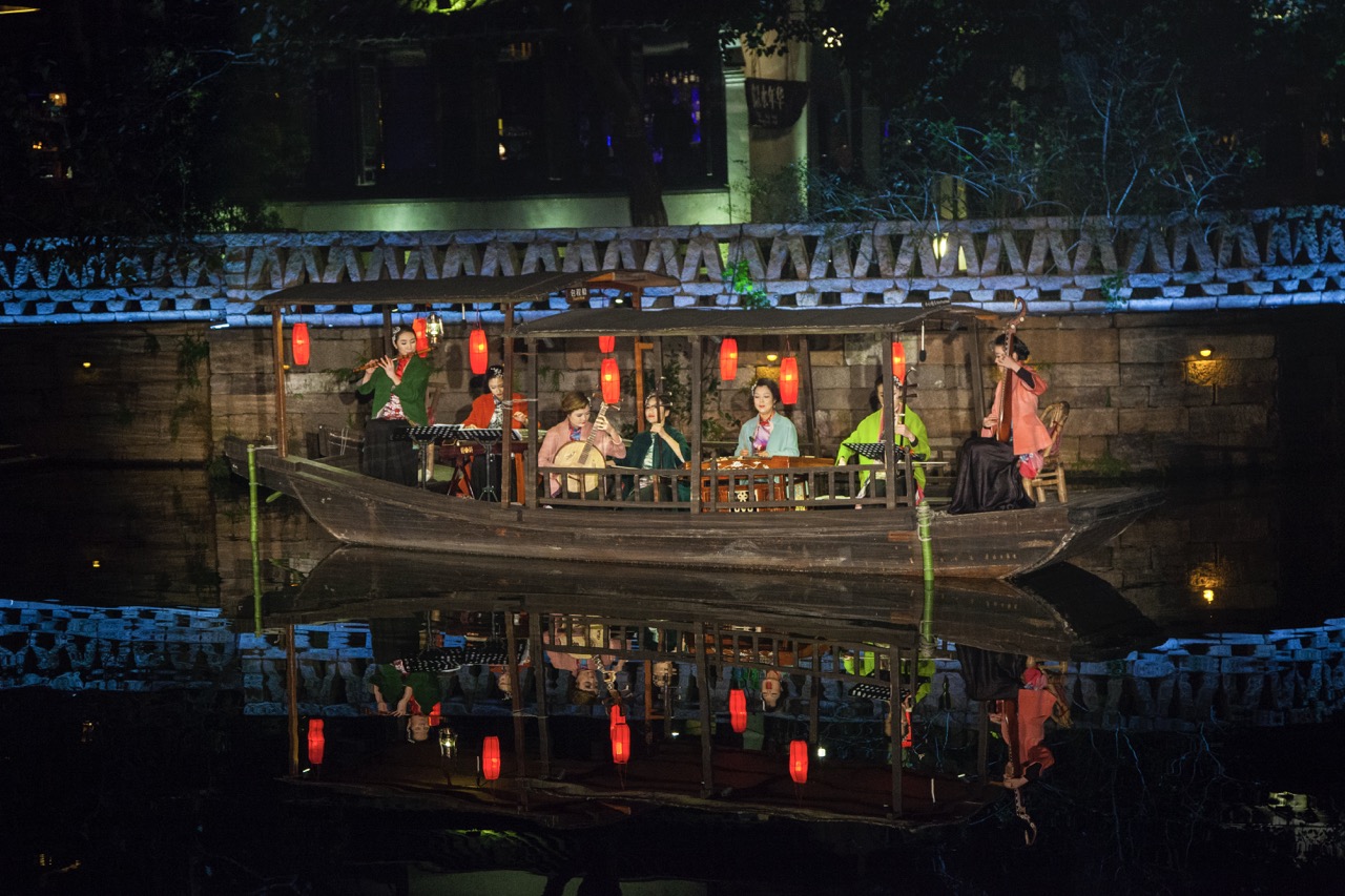 Traditional Chinese musicians performing on a wooden boat with red lanterns at night, reflected on a calm canal