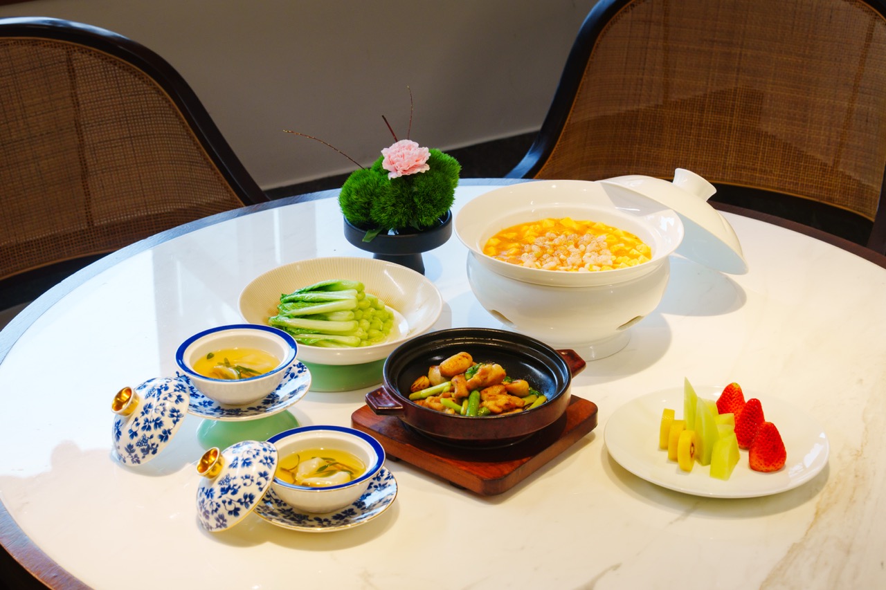 Delicate assorted Jiangnan dishes including seafood soup, braised clay pot dish, seasonal greens and fresh fruit platter on white marble tabletop.