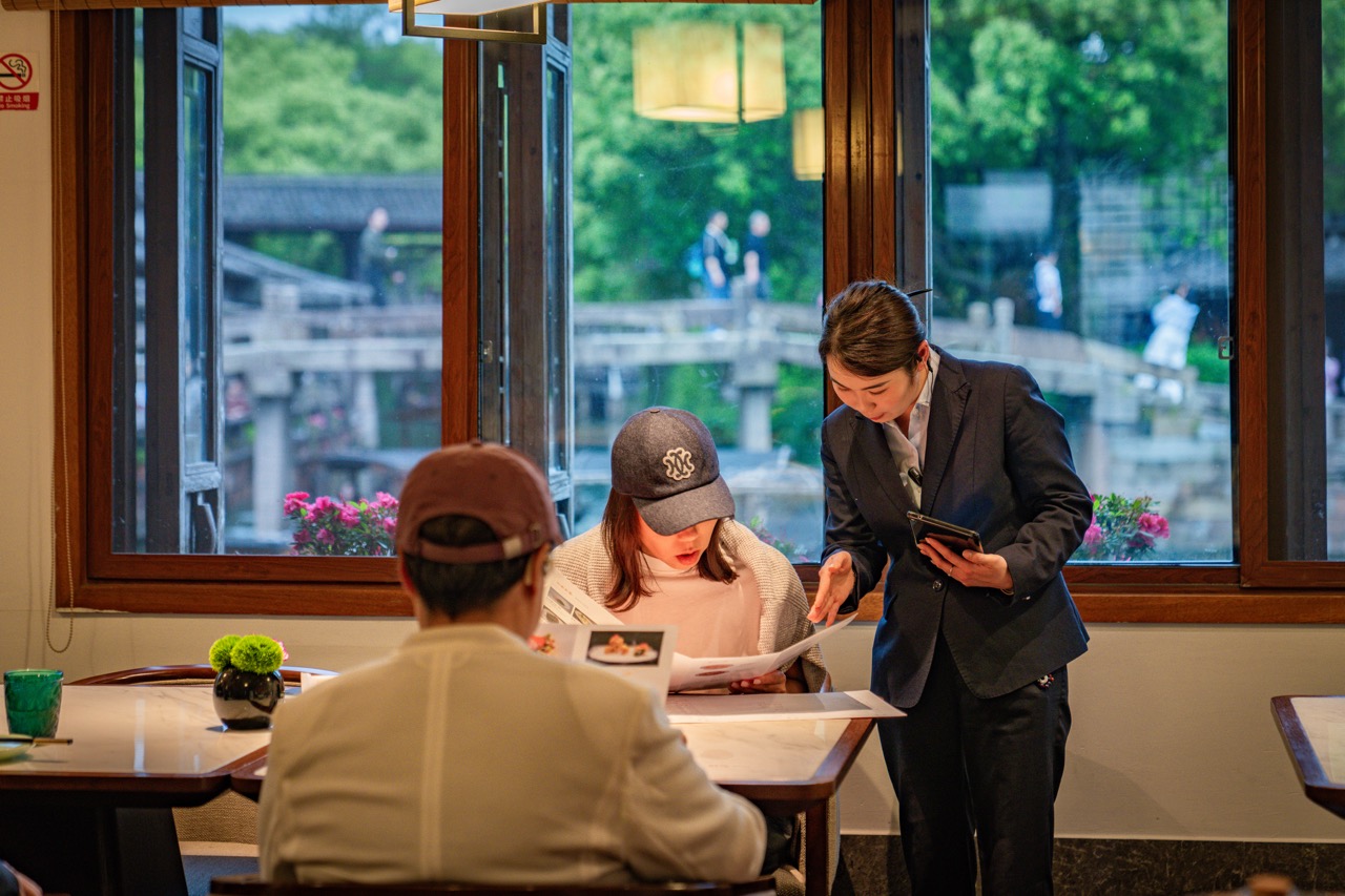 Professional female service staff holding tablet introducing menu to diners beside large wooden window overlooking Wuzhen canal stone bridge and green scenery.