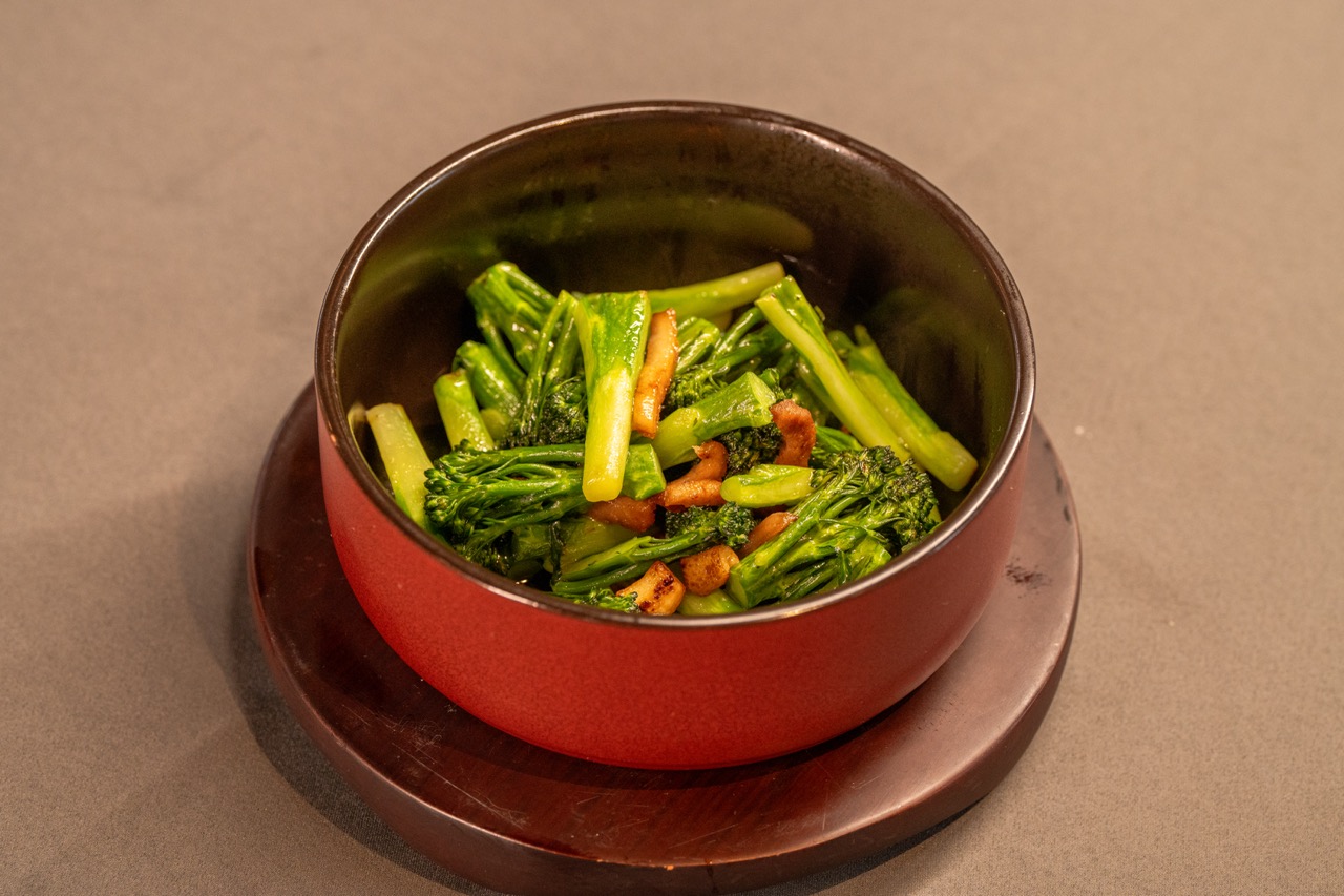 Crisp green broccolini stir-fried with sliced bacon in red bowl.