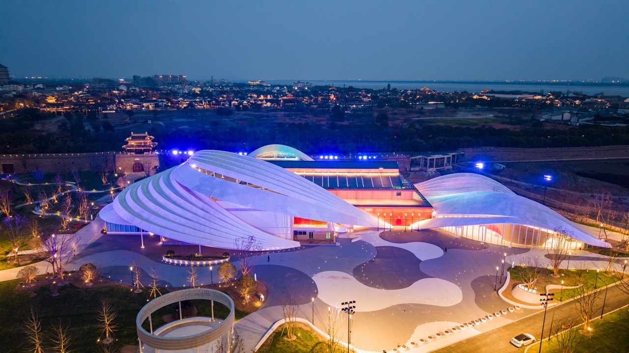 Overhead night view of Yanguan Grand Theatre of Nations with flowing curved white roof lighting, ancient town and wide lake stretching in background