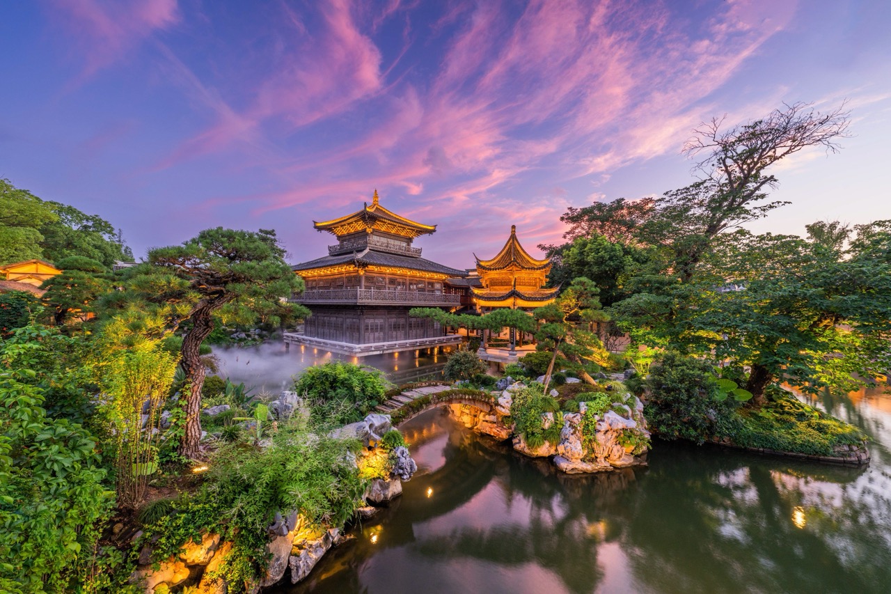 Lit traditional pavilions reflect on calm garden water under pink dusk sky.