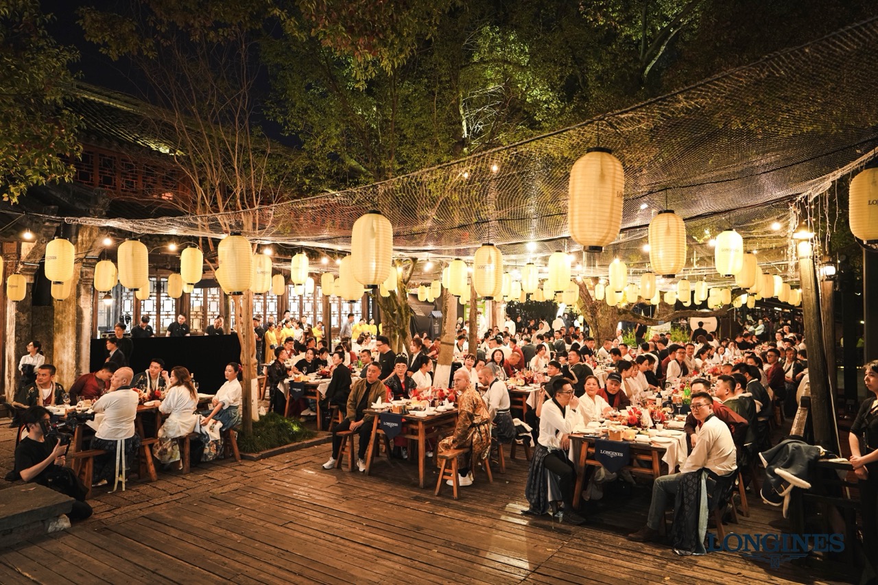 Outdoor brand dinner event of Longines in Wuzhen, rows of warm paper lanterns hanging over wooden dining tables with numerous guests attending at night.