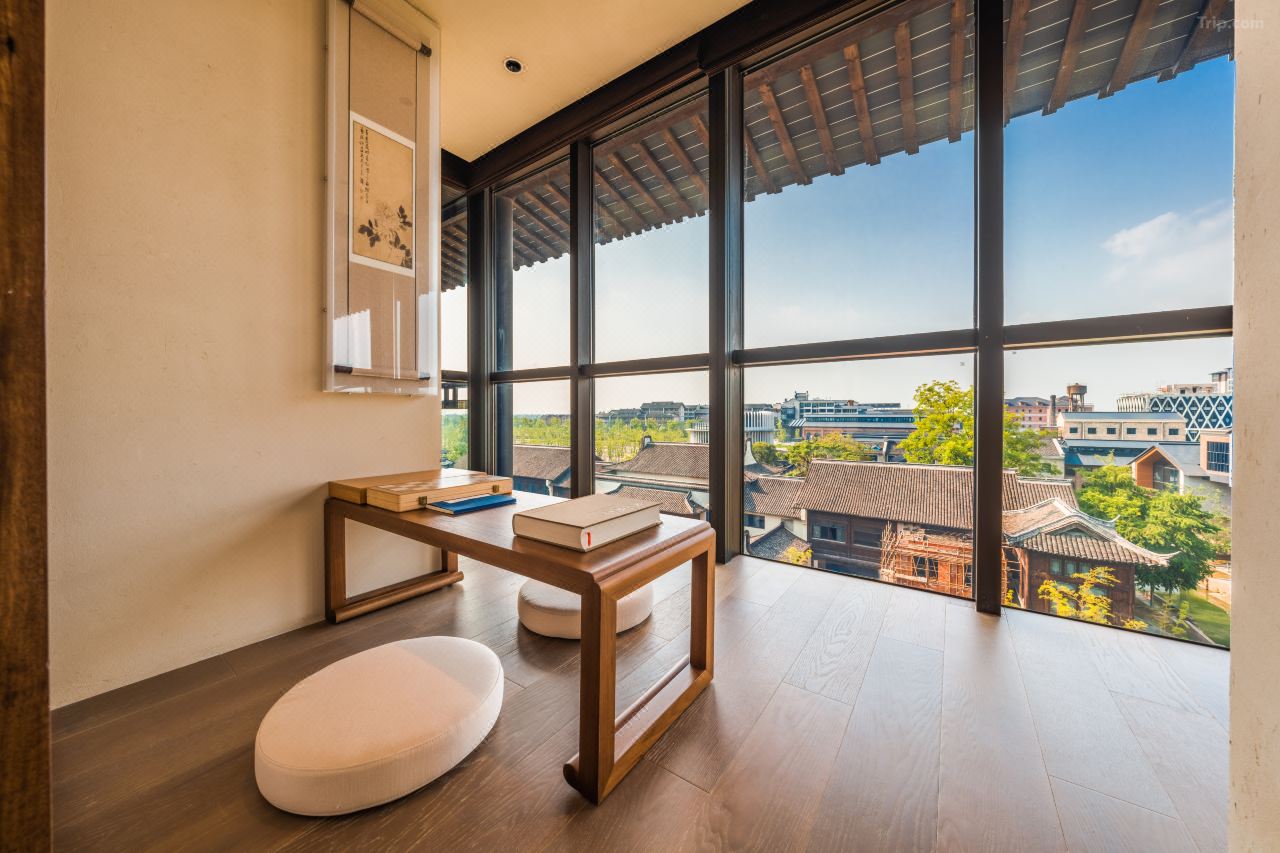 Low wooden desk with floor cushions, floor-to-ceiling glass framing tiled ancient water town rooftops.