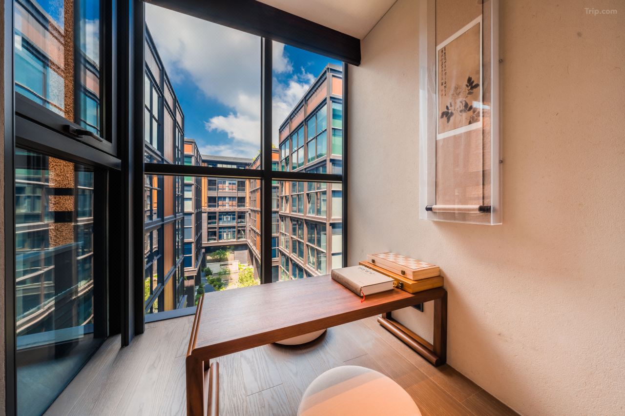Warm wood writing table beside floor-to-ceiling window, framed Chinese painting hanging on plaster wall.