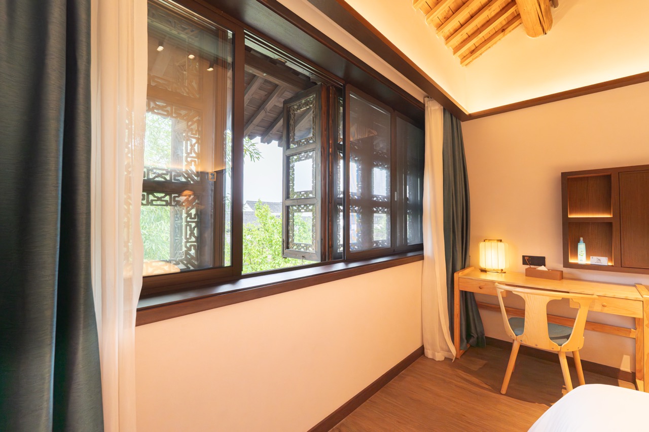 Large antique-style carved window with layered curtains, wooden desk and view of courtyard greenery.