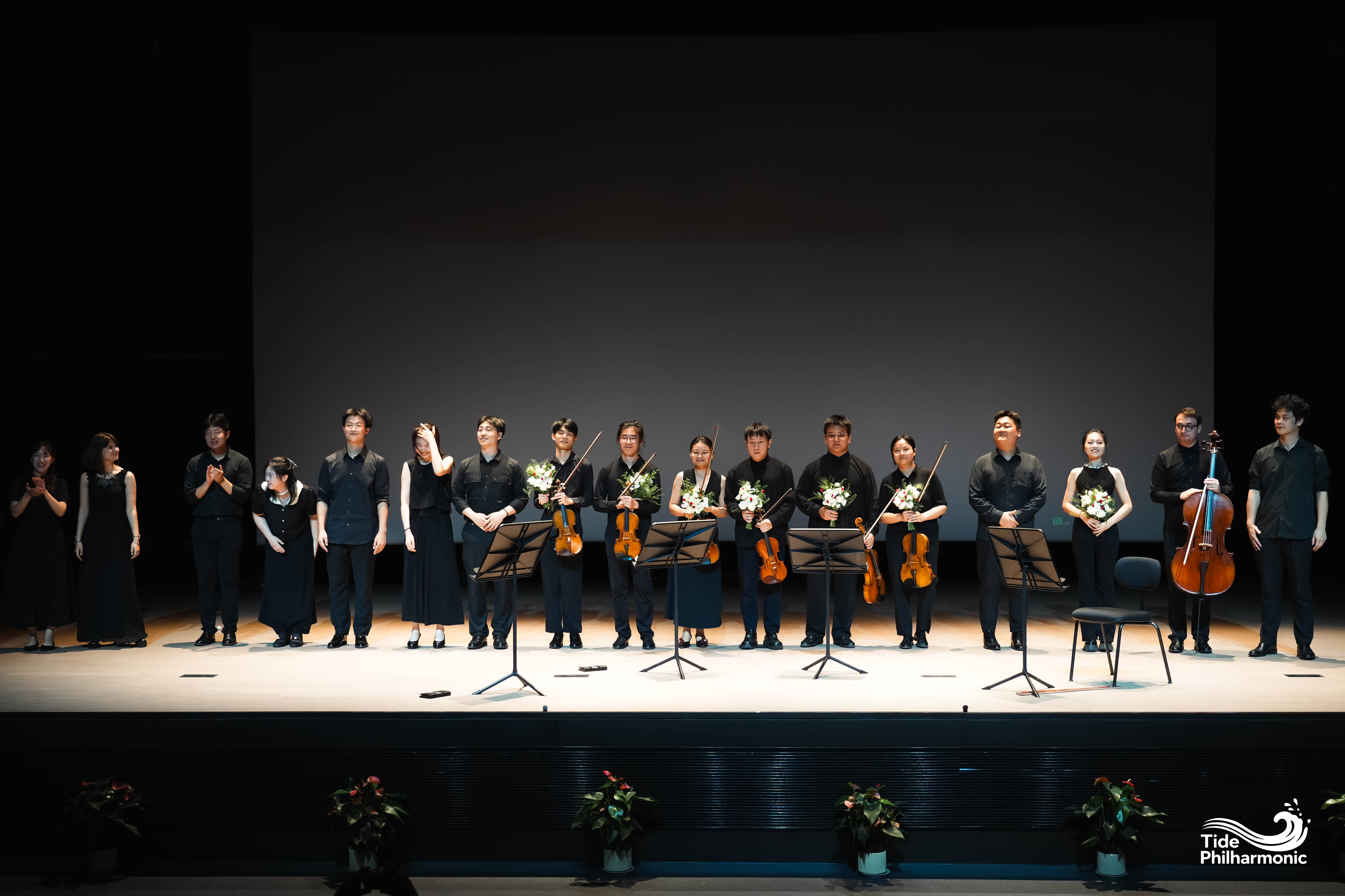 String musicians standing in line on stage holding bouquets after performance, Tide Philharmonic logo at bottom right.