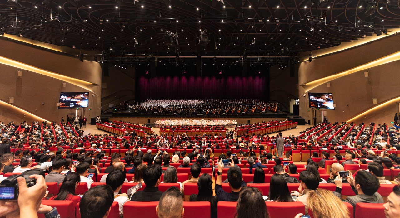 Wide view of fully occupied red-seat auditorium facing symphony stage, audience capturing show with phones.