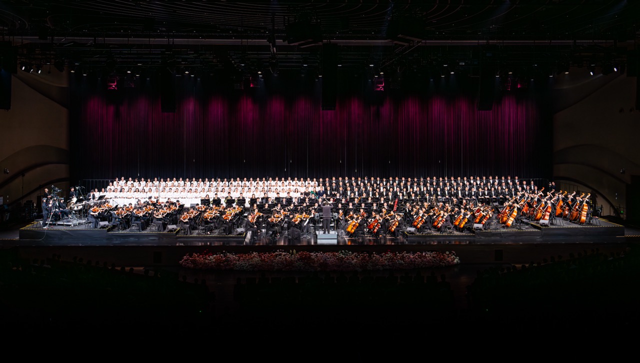 Panoramic shot of full Tide Philharmonic orchestra plus large choir lined up on main stage under dark theatre lighting.