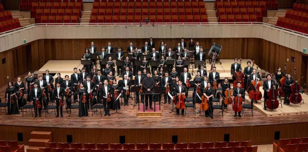 Formal group portrait of Tide Philharmonic Orchestra with conductor on wood-paneled opera stage.
