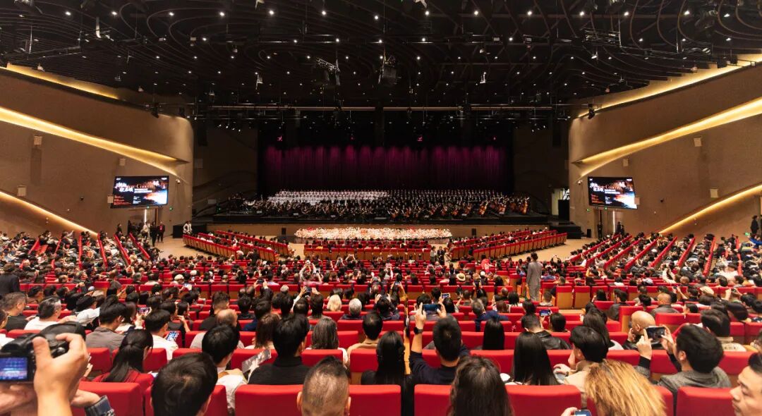 Packed auditorium view of the premiere performance of Jiaxing Tide Philharmonic Orchestra at Wanguo Opera House, full orchestra and choir on stage with audience taking photos.