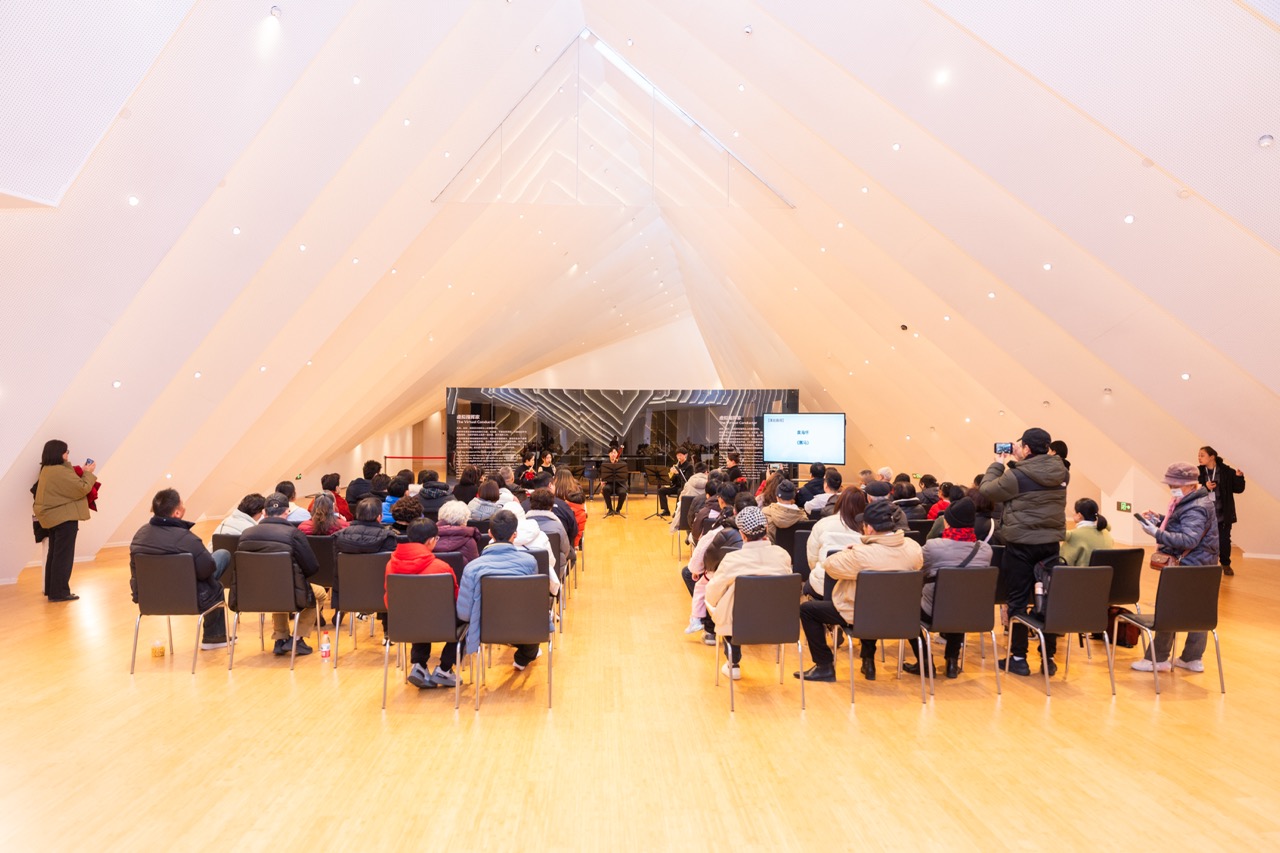 Audience seated in bright triangular-ceiling recital hall watching chamber musicians perform on small stage.