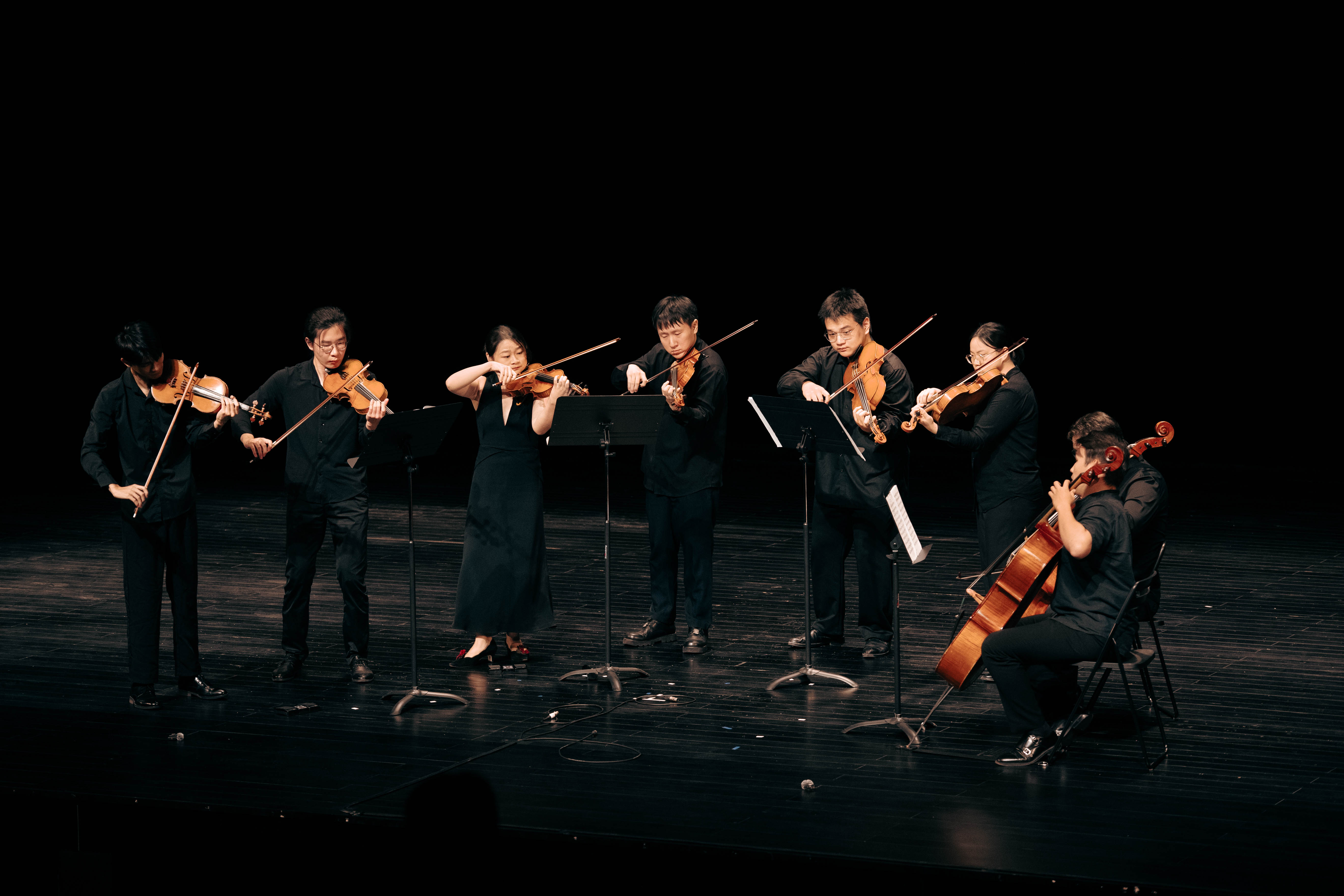 Seven-piece string group playing violins, viola and cello on minimalist black stage.