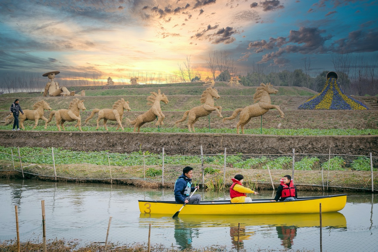 Visitors paddling bright yellow canoe, terraced farmland lined with hand-woven straw horse art under dusk sky.