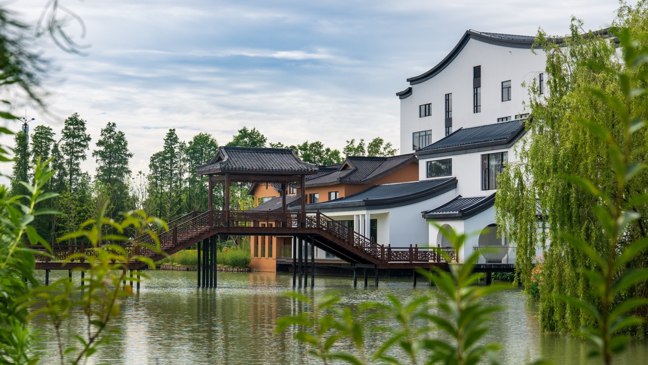 White-and-black Huizhou architecture connected by wooden covered bridge, willow and fresh green foliage framing calm water.