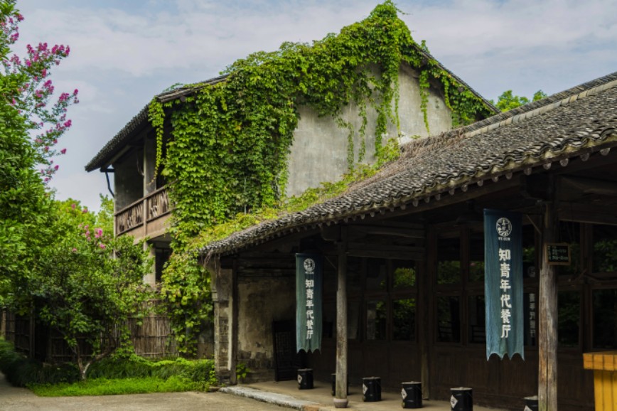 Old Jiangnan timber house covered with ivy, hanging blue vertical banners printed with restaurant name, rural ancient building scenery.