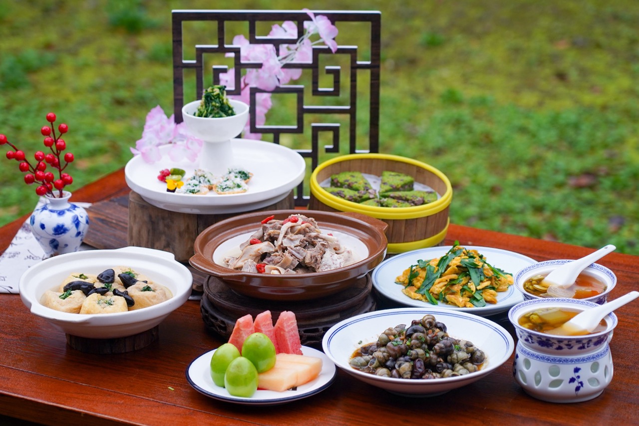 Full spread of local Wucun specialties including braised mutton, stir-fried river snails, seasonal greens, tofu casserole and fruit platter arranged on wooden table outdoors.