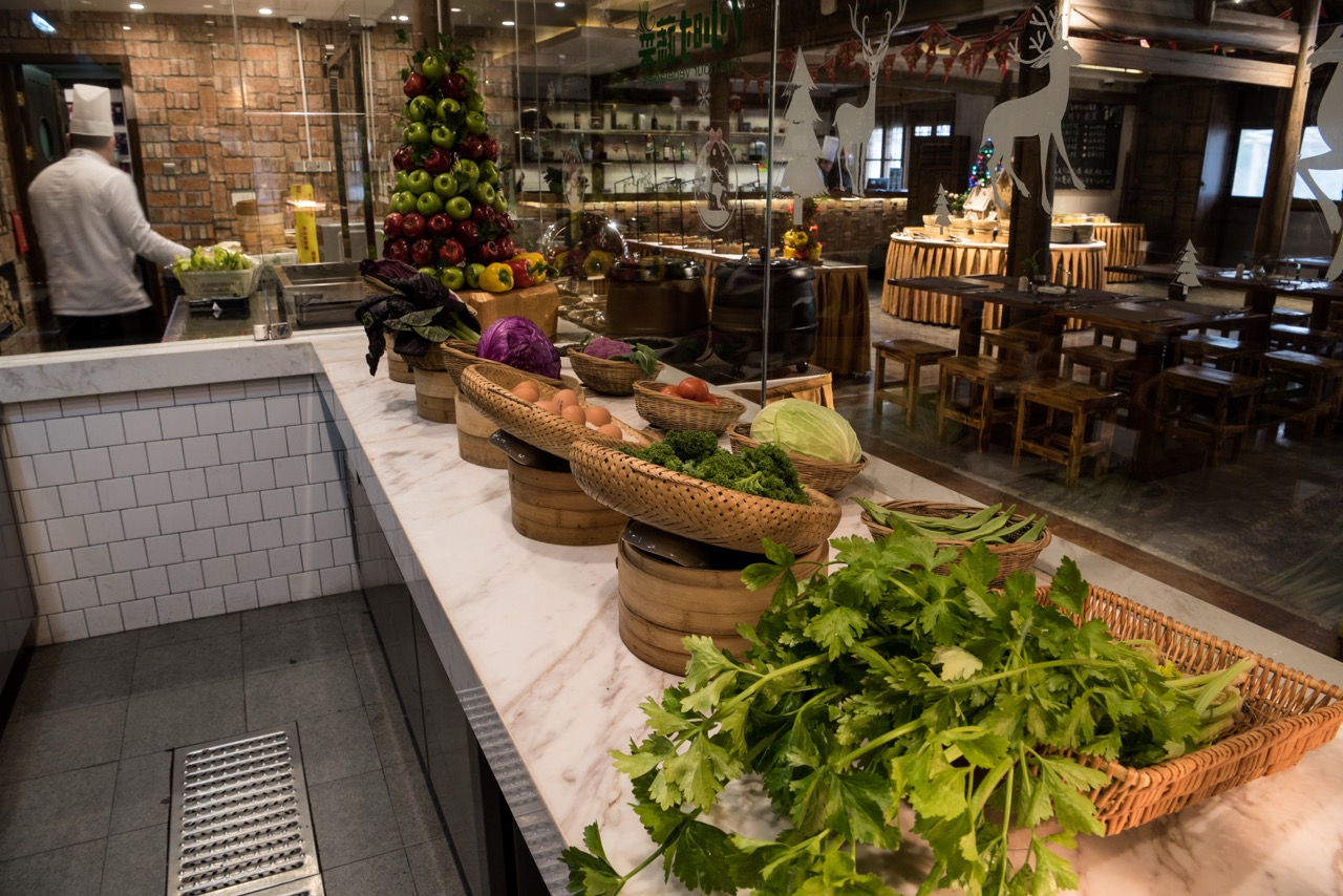 Open kitchen fresh vegetable display counter alt：Marble serving counter stocked with seasonal local greens, bamboo baskets and bamboo steamers, transparent glass viewing kitchen area.