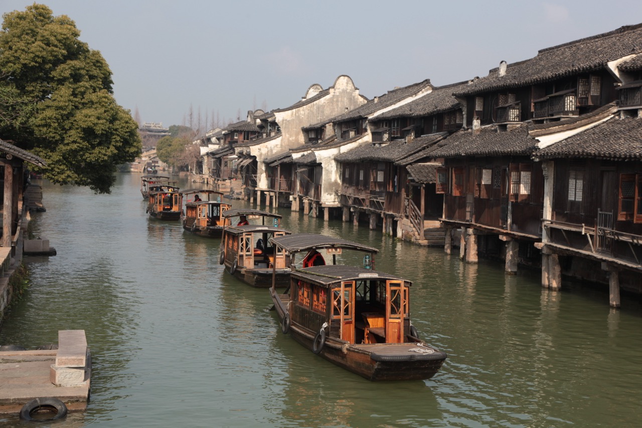 Row of covered wooden sightseeing boats docked beside stilted ancient dwellings on Wuzhen canal.