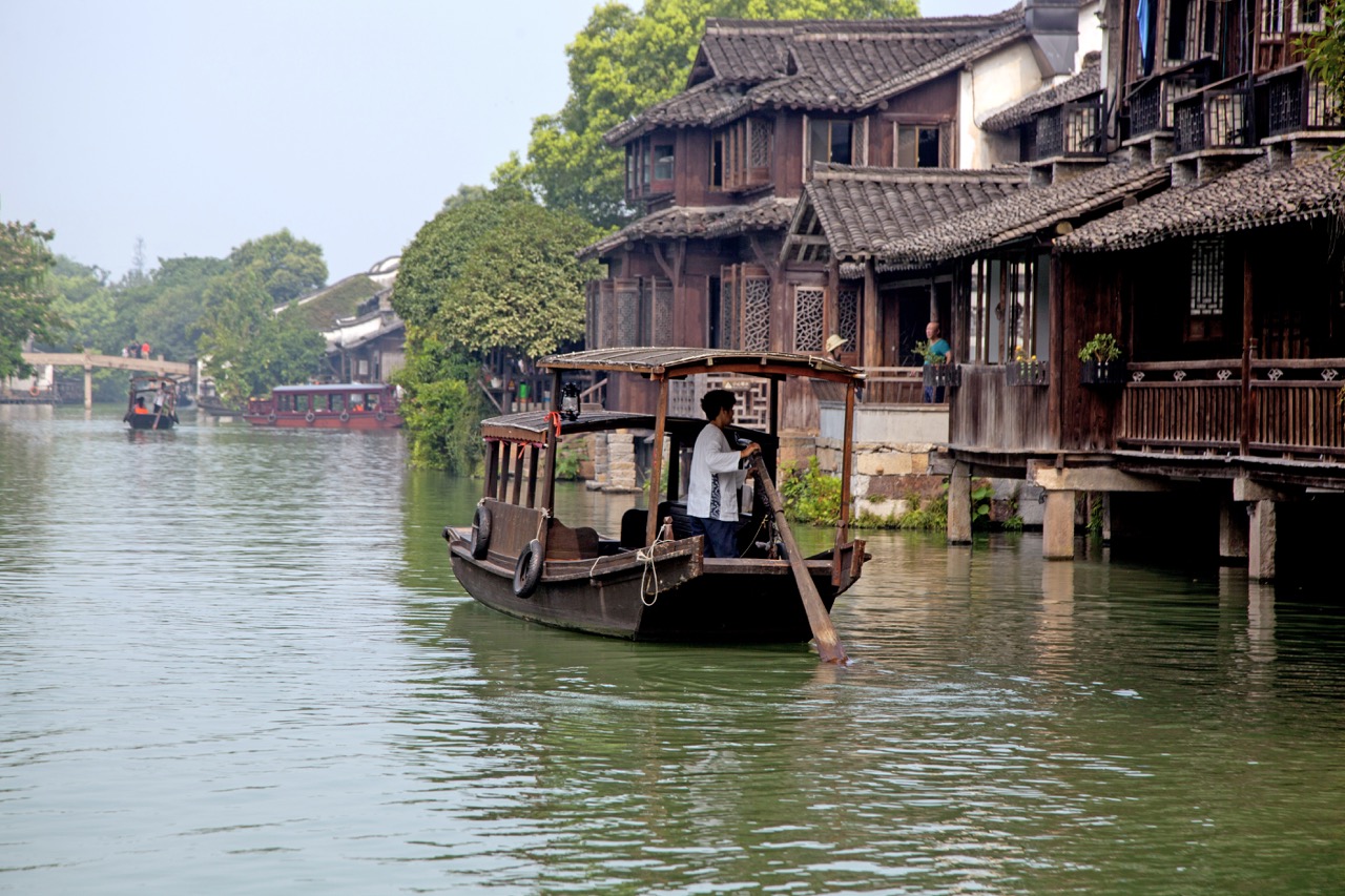 Local boatman standing at stern, poling wooden sightseeing boat past waterside courtyard houses in Wuzhen.