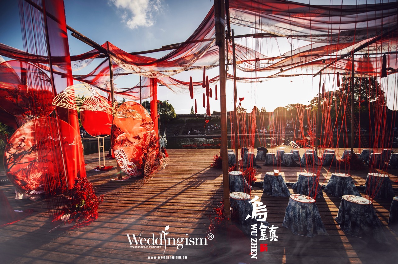 Outdoor waterfront wedding stage decorated with red silk fabric, Chinese landscape art installations and hanging red tassels for traditional wedding ceremony in Wuzhen water town.