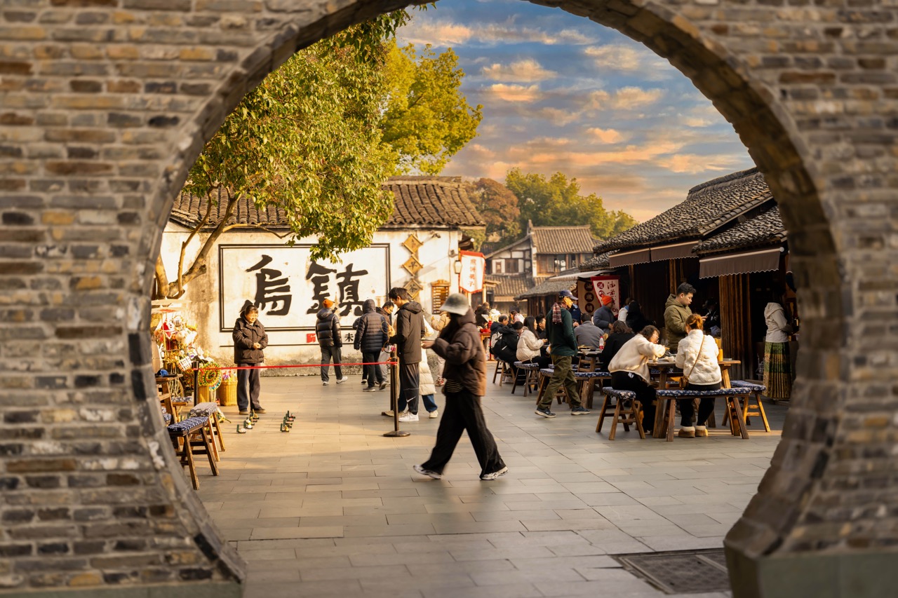 Scene of bustling morning teahouse street framed by round stone arch, big "Wuzhen" wall mural.