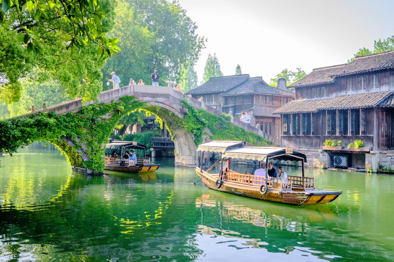 Traditional wooden covered boats cruising under ivy-covered stone arch bridge, ancient water town scenery for corporate group team building activity in Wuzhen.