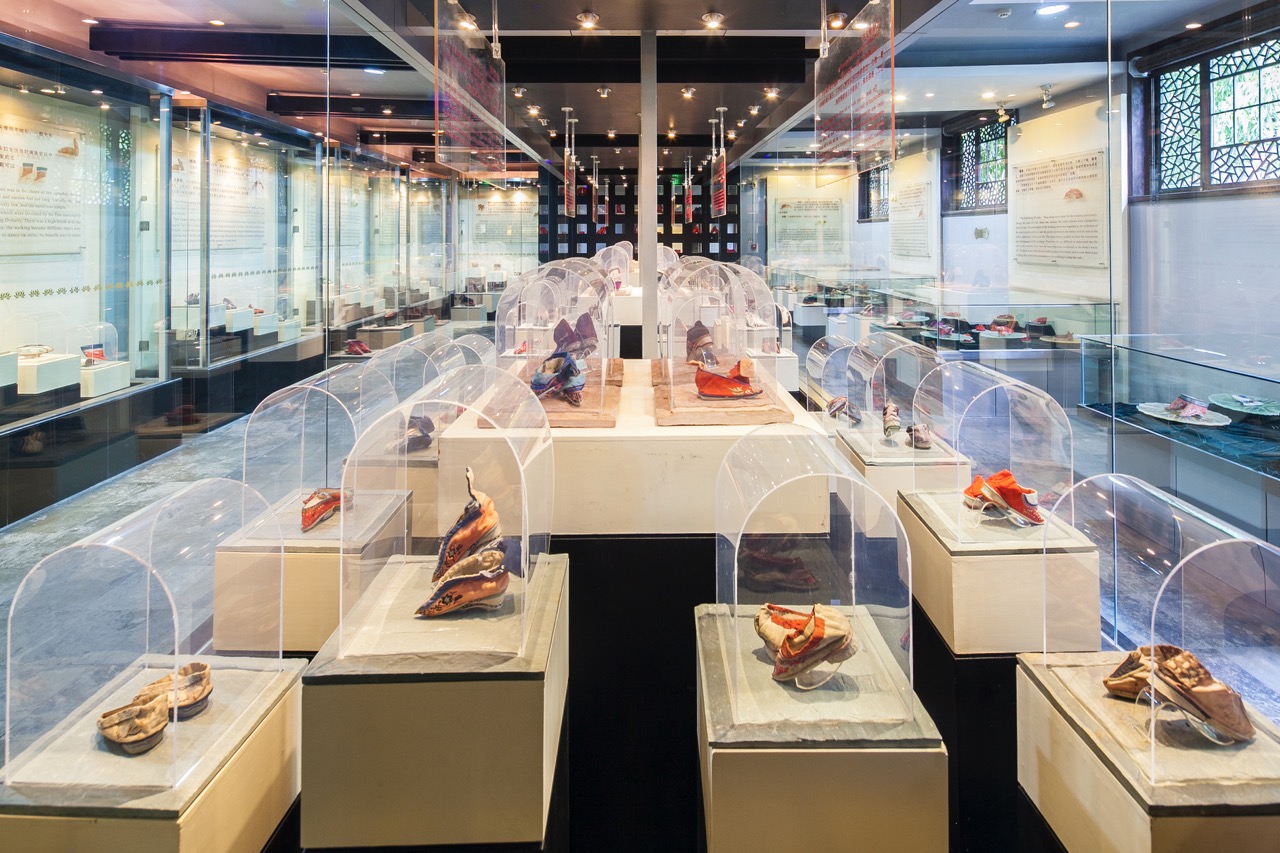 Glass display stands showcasing antique embroidered lotus shoes inside cultural museum.