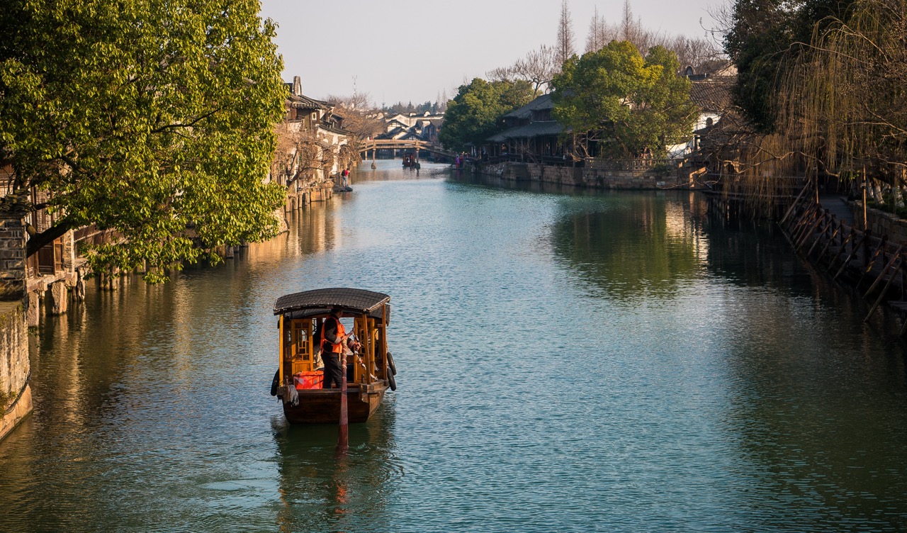 Single hand-rowed wooden boat floating on calm canal water, flanked by ancient waterside houses and lush green trees in Wuzhen water town.