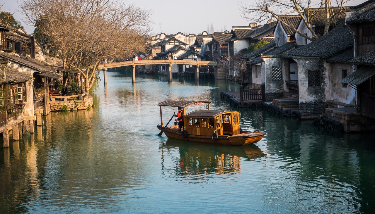 Classic wooden sightseeing boat sailing through water town canal, stone foot bridge and old Jiangnan architecture in background.