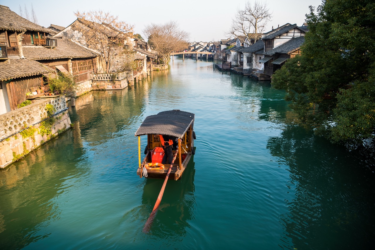 High-angle shot of covered wooden boat gliding down quiet waterway lined with historic timber buildings in Wuzhen.