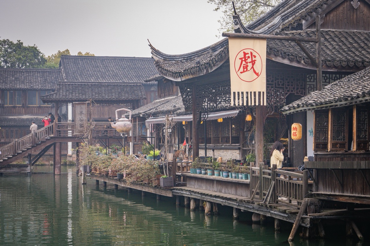 Stilted wooden teahouse along canal, hanging red Chinese opera flag and giant teapot decor.