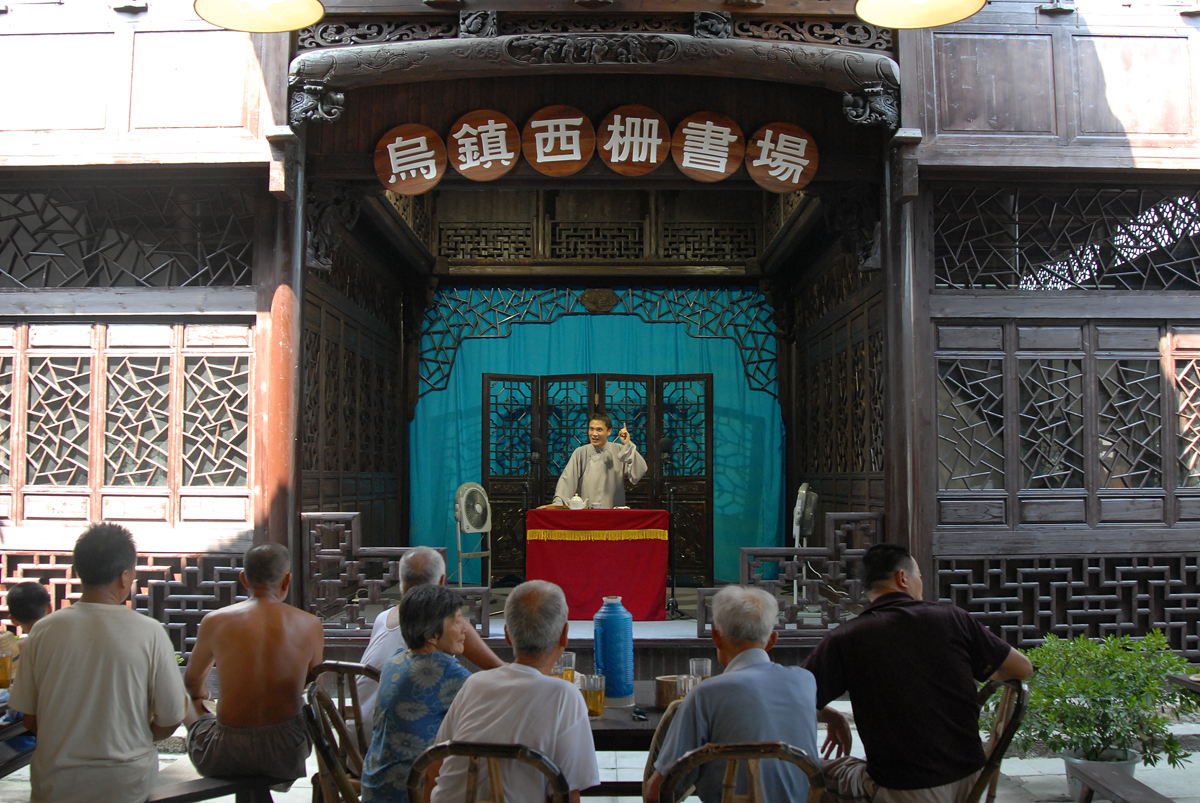 Folk storyteller performing at Wuzhen Xizha Book House, local audience seated watching traditional pingtan show.