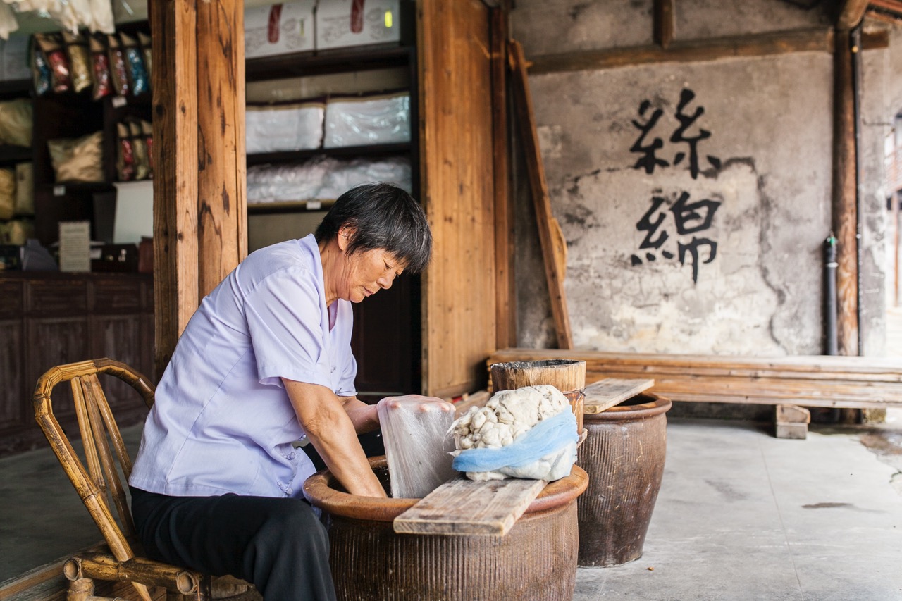 Elderly craftswoman hand-making silk floss beside clay vat at Yida Silk House, wall painted with Chinese characters "silk floss".