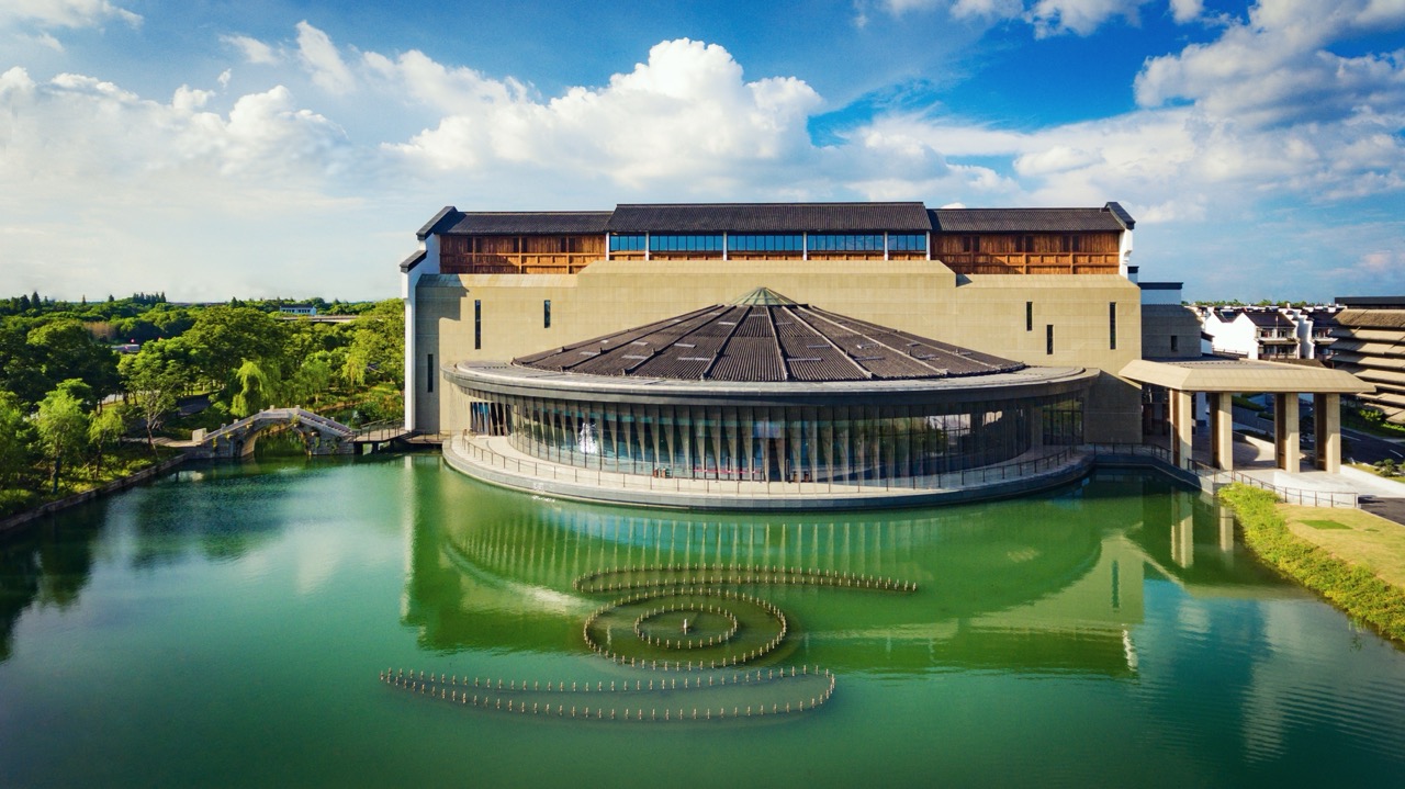 Aerial shot of Yunzhou Guest Center in Puyuan Fashion Resort, modern Jiangnan-style architecture mirrored in green lake water with spiral fountain installation.