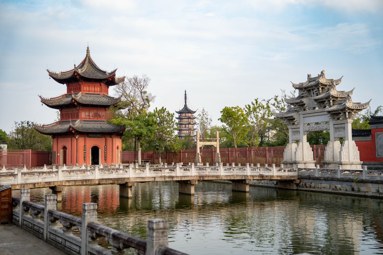 Red octagonal pavilion, stone memorial arch and Bailian Pagoda reflected in canal water.