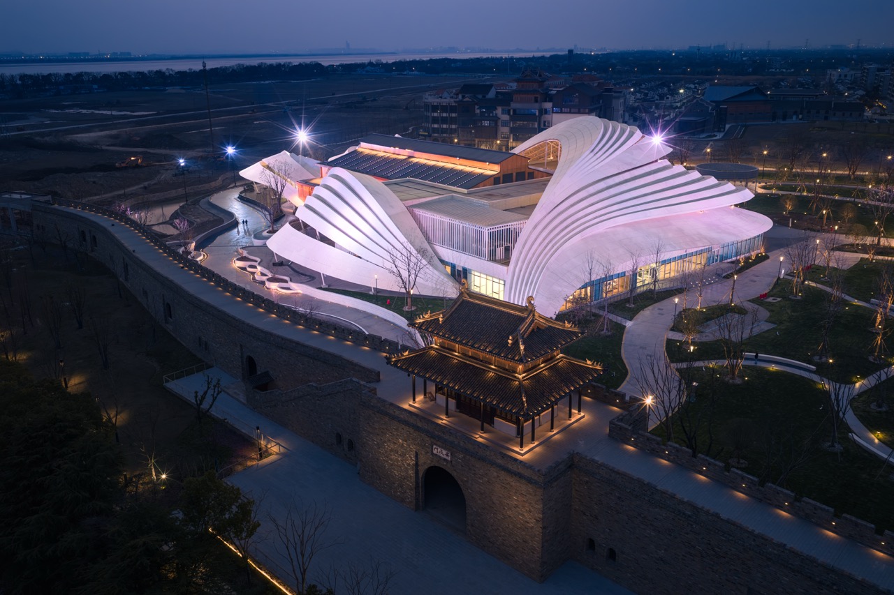 Historic stone city wall pavilion in foreground, modern wave-shaped opera theatre illuminated behind it at dusk.