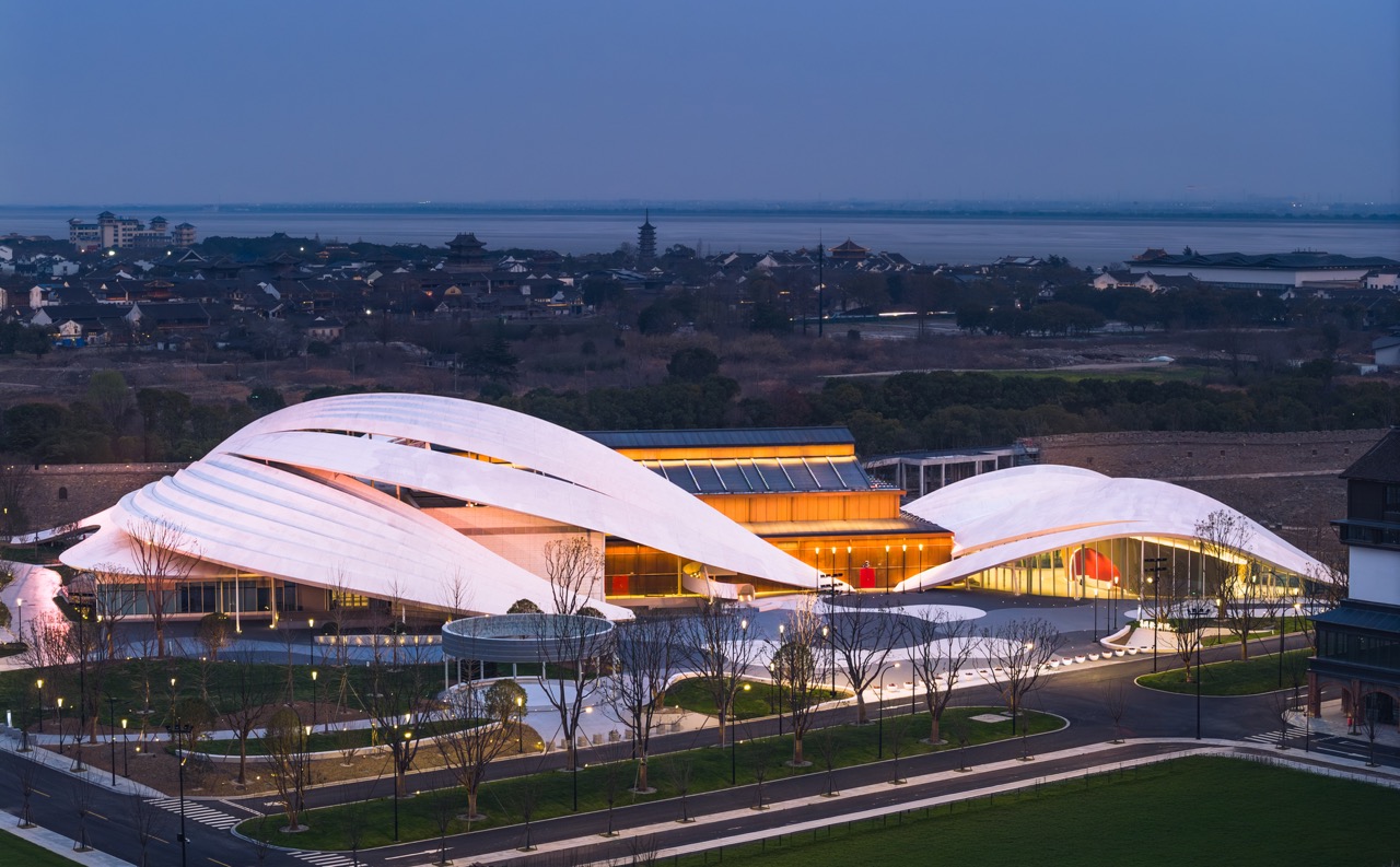 Curved white wave-shaped opera building at twilight, Qiantang river and Bailian Pagoda visible far in background.