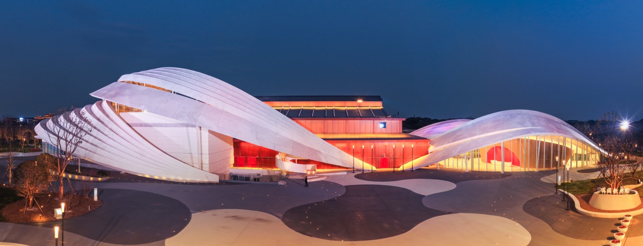 Wide front shot of flowing ripple roof opera building with warm interior lighting and abstract fluid plaza pavement.