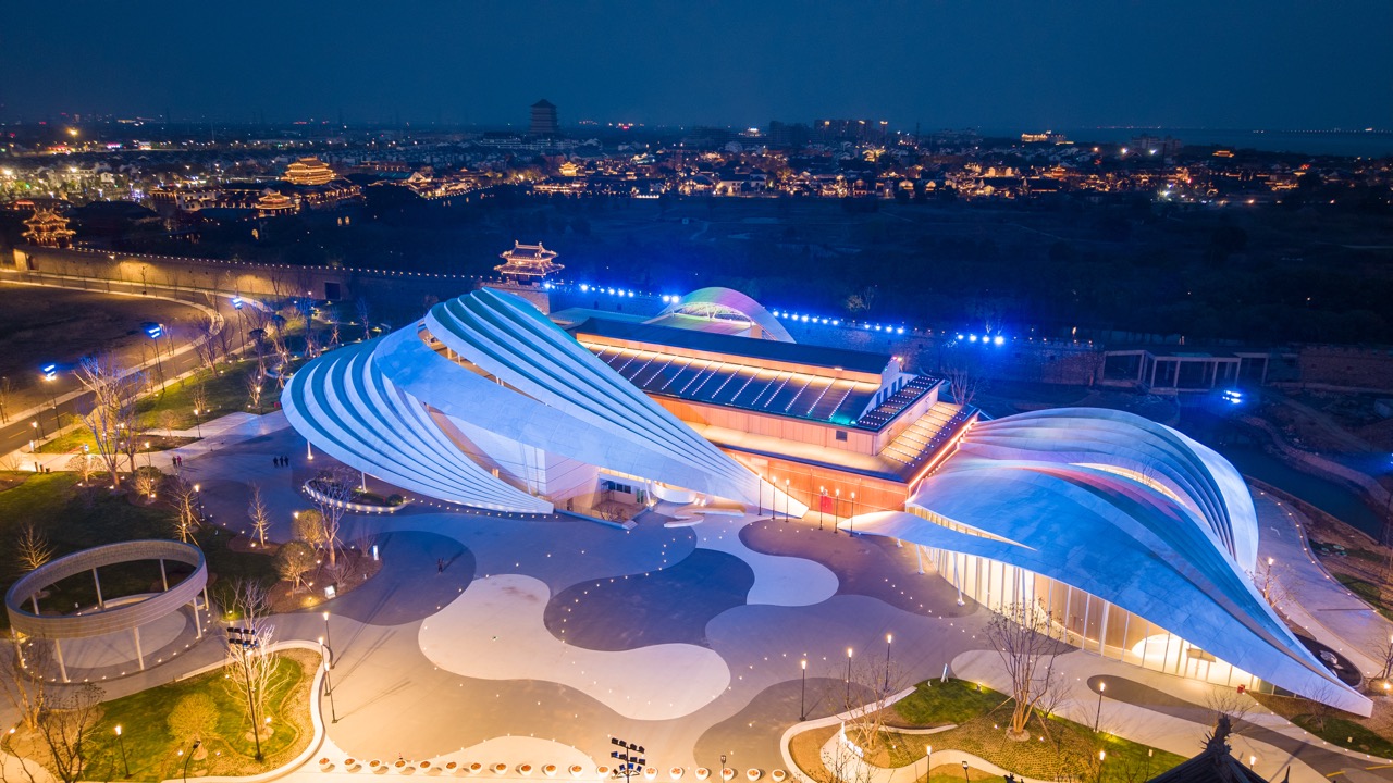 Overhead shot of wave-form opera hall with colorful facade lighting, ancient town buildings stretching behind.