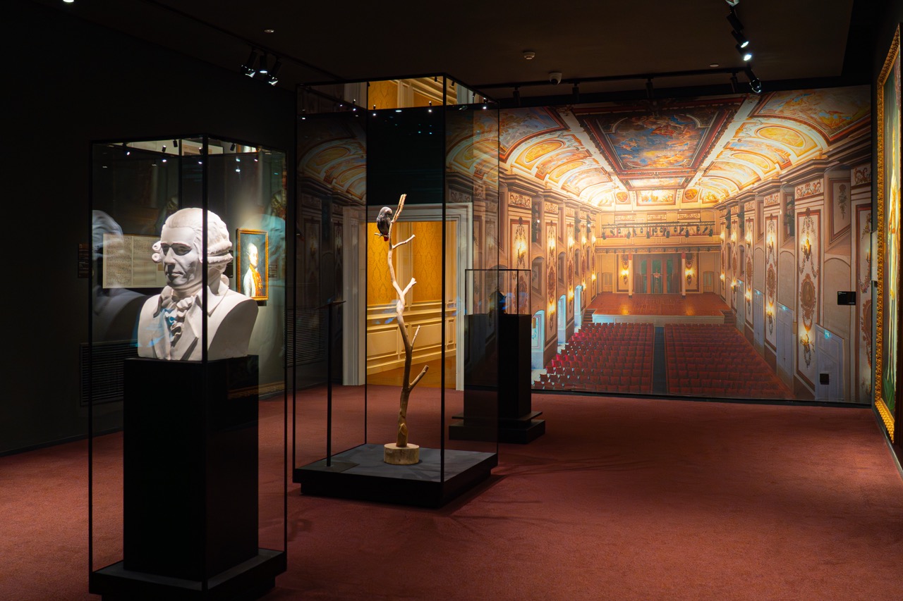 Narrow warm-toned corridor lined with glowing pyramid display cases holding classical musician bust sculptures.