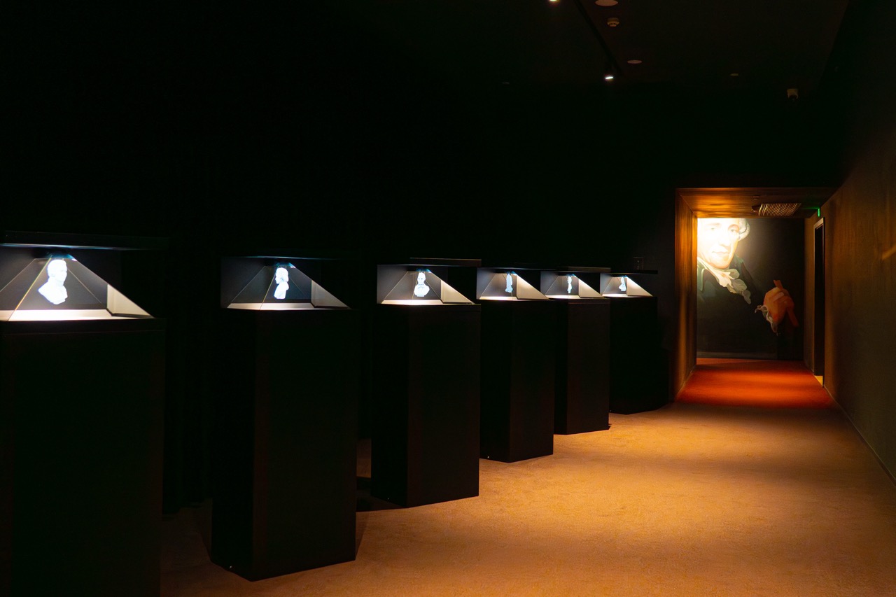 Dark exhibition room with glass display cases holding composer bust sculptures, large mural of classic opera hall on rear wall.